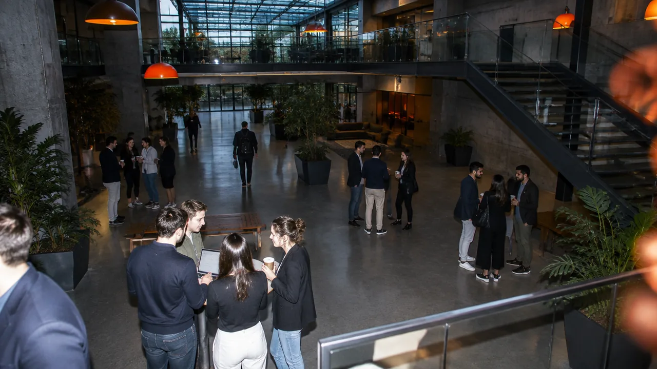 View from a mezzanine into a concrete and glass startup hub atrium where small groups of founders, mentors and investors talk during a session break under orange pendant lights