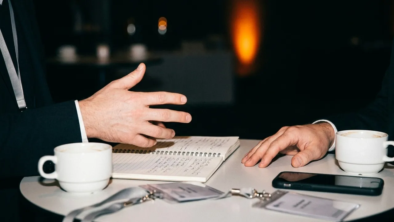 Close-up of two people's hands during a one-to-one meeting at a small conference table, with badges, notebooks and coffee cups, lit by camera flash against a dark event hall