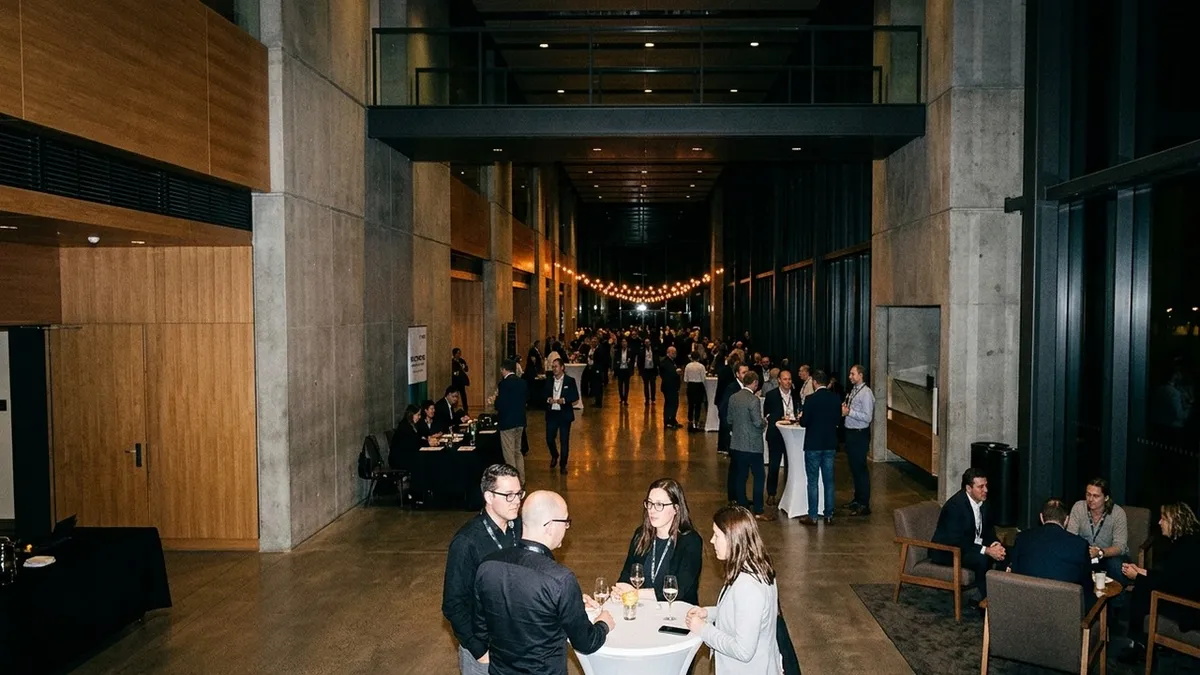 Evening networking reception in a large conference foyer: attendees talking at standing tables and in lounge corners, with string lights above the crowd in the background.